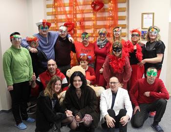 Uczestnicy oraz kadra ubrani na czerwono w maskach karnawałowych pozujący do zdjęcia grupowego na tle czerwon